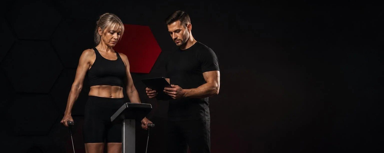 Suivre ses progrès : composition corporelle et récupération Femme de 50 ans sur un analyseur de composition corporelle pendant que son coach consulte les résultats